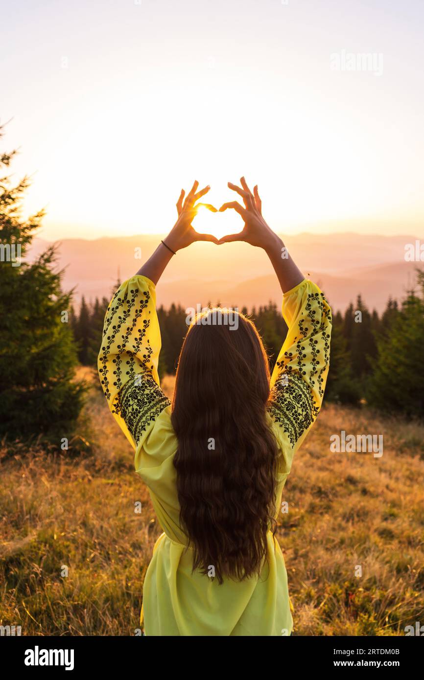 A girl holds her hands in the shape of a heart at dawn in the mountains. A girl in national Ukrainian clothes. Romance in the mountains. Stock Photo