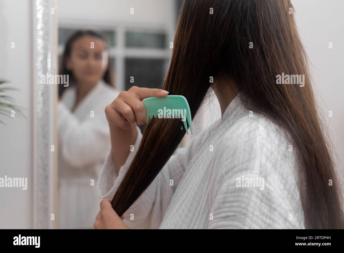 Woman combing long straight chestnut hair looking in mirror in bathroom hairstyling with comb Stock Photo