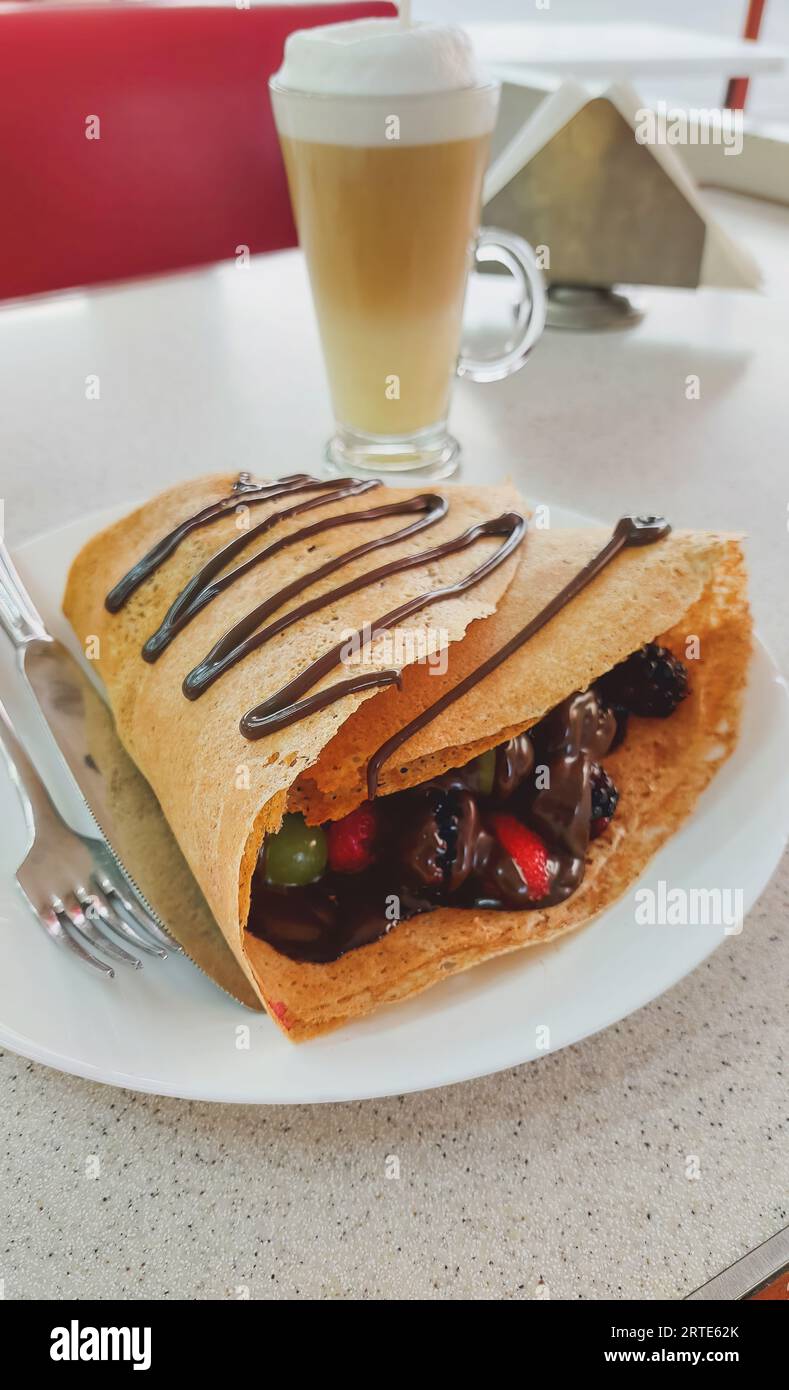 Vertical view of a red fruit crepe with chocolate, containing strawberries, blackberries, grapes and melted chocolate, with a cappuccino coffee in the Stock Photo