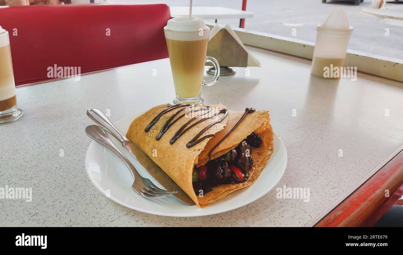 Horizontal view of a red fruit crepe with chocolate, containing strawberries, blackberries, grapes and melted chocolate, with a cappuccino coffee in t Stock Photo