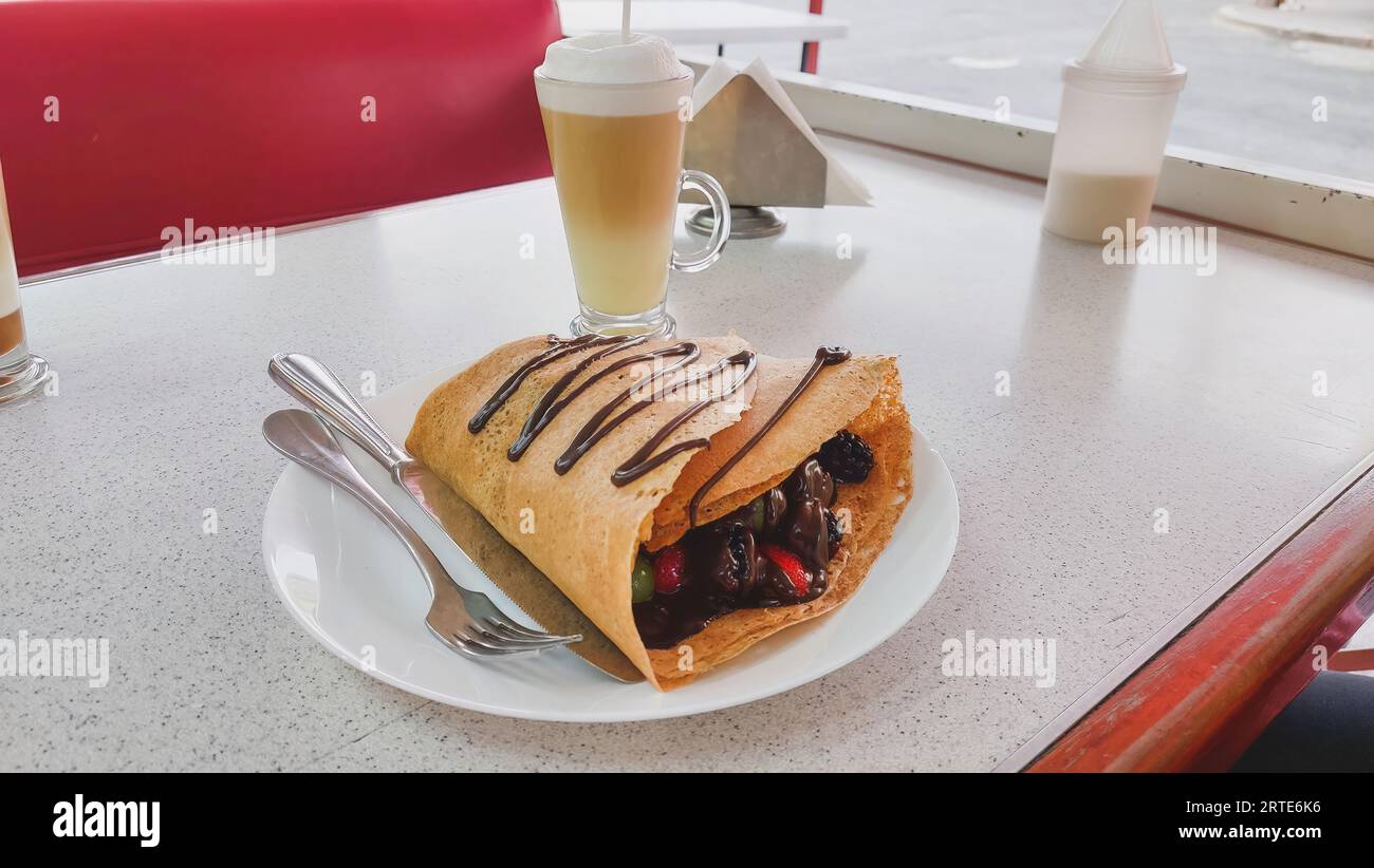 Horizontal view of a red fruit crepe with chocolate, containing strawberries, blackberries, grapes and melted chocolate, with a cappuccino coffee in t Stock Photo