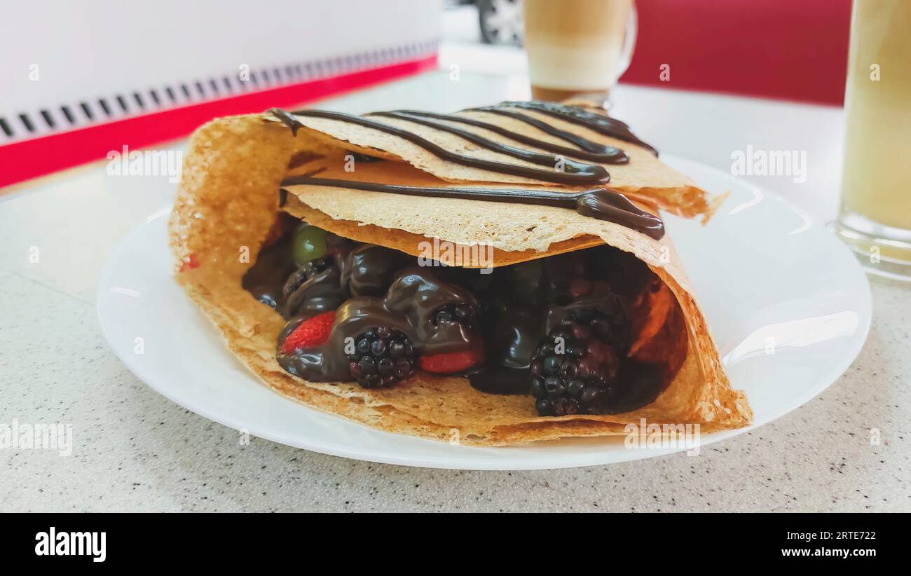 Close-up of a red fruit crepe with chocolate, containing strawberries, blackberries, grapes and melted chocolate, with a cappuccino coffee in the back Stock Photo