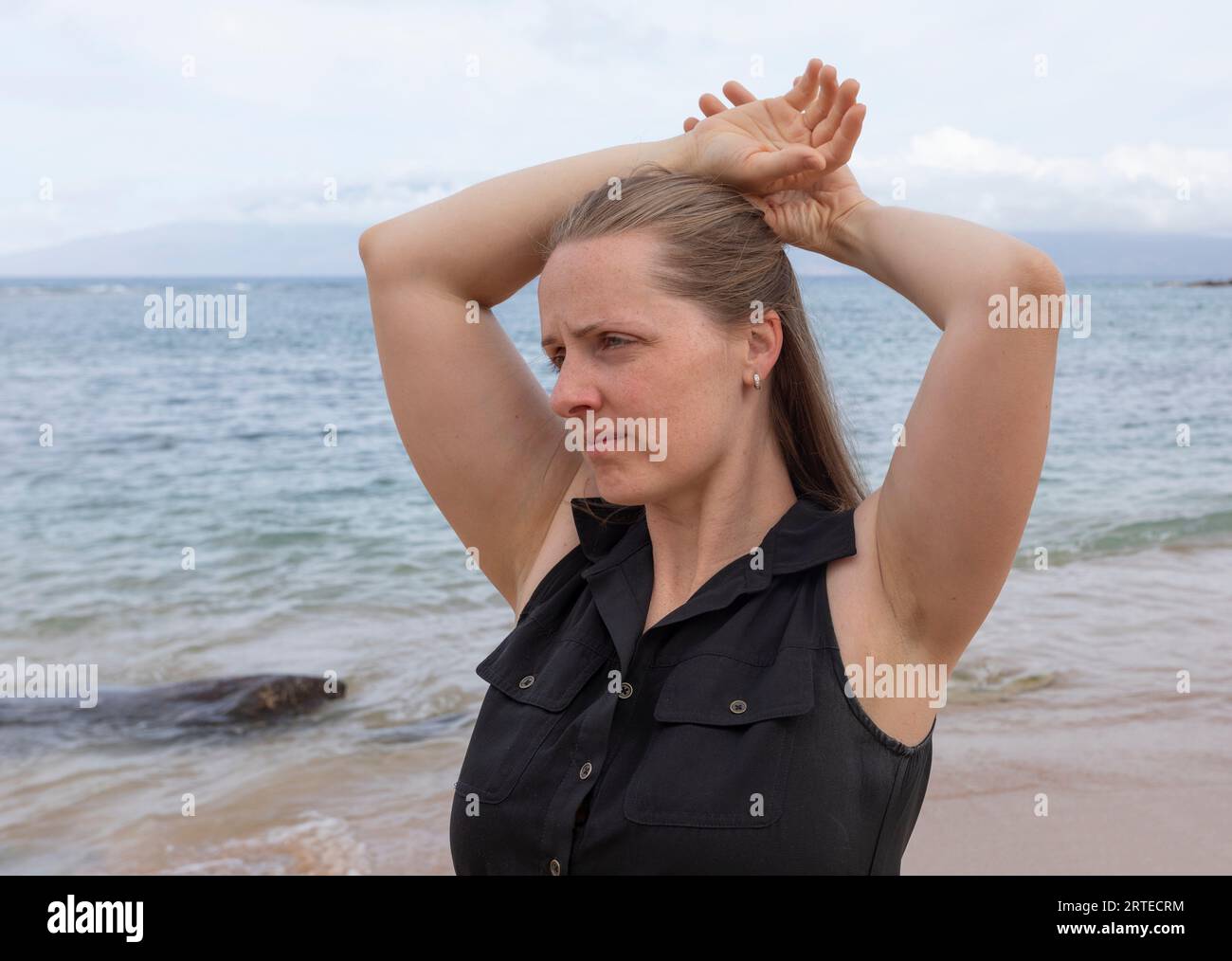 Portrait of a woman standing on the shore with arms behind head, looking pensively at the ocean from Kapalua Beach in West Maui Stock Photo