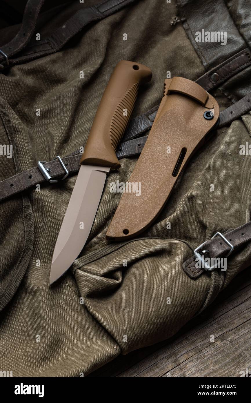 Modern knife with rubberized handle and plastic sheath. Cold steel lies on a vintage hiking backpack. Wooden background. Stock Photo