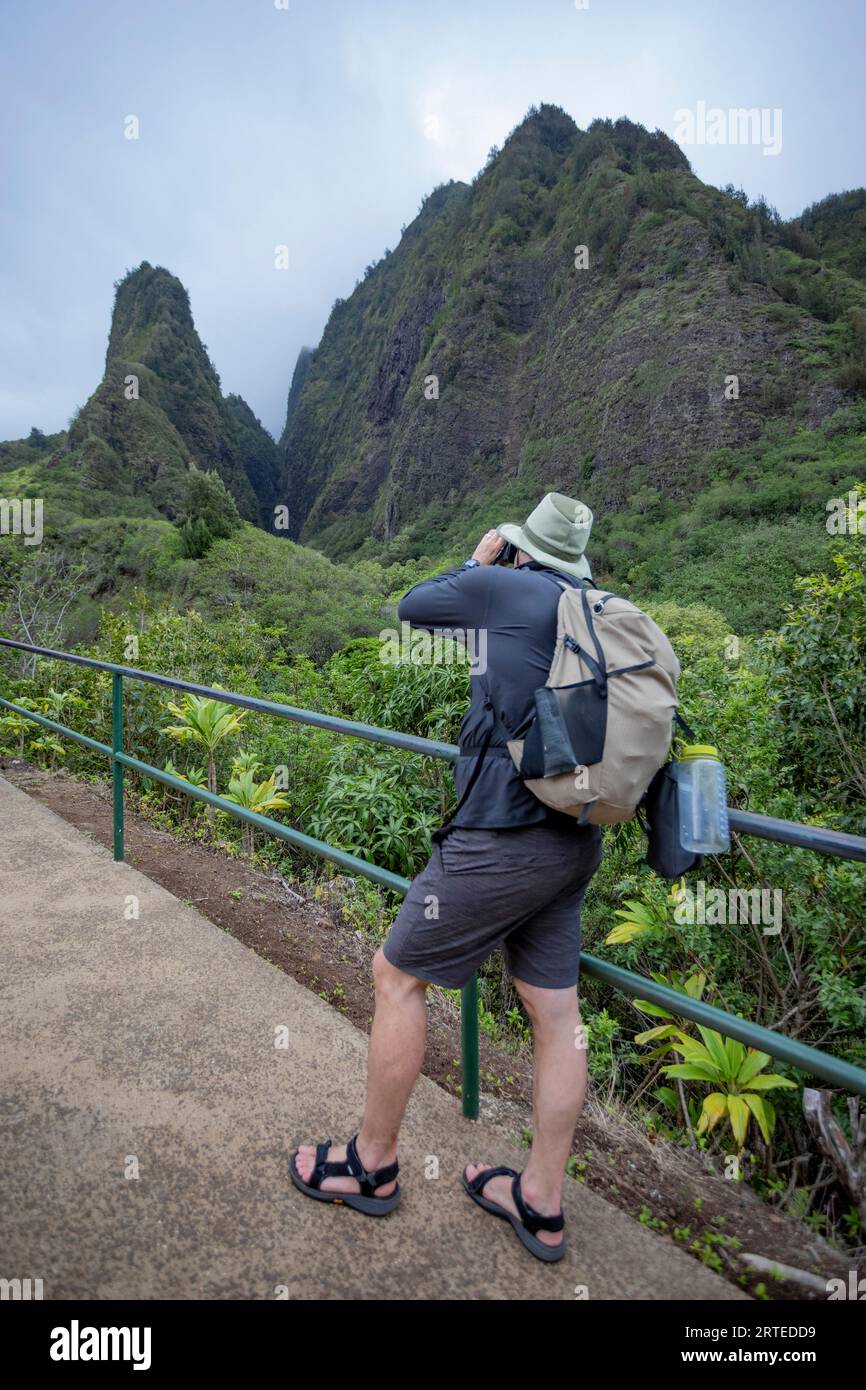 View taken from behind of a man standing on a walkway photographing the view of the Kuka‘emoku, Iao Needle in the lush, Iao Valley in Central Maui Stock Photo