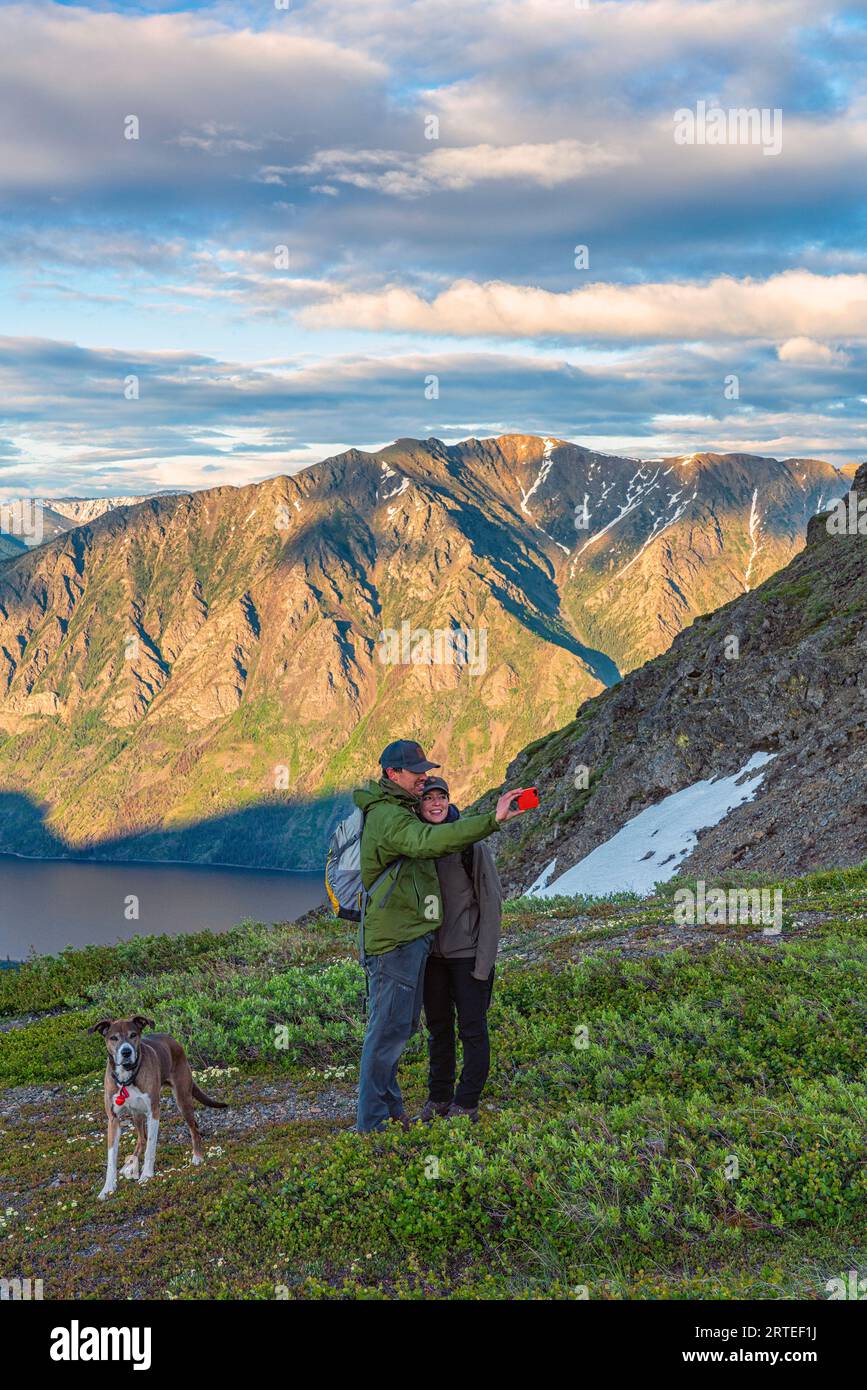 Couple taking a picture of themselves on top of a mountain at twilight with beautiful scenic backgound, while their dog stands beside them looking ... Stock Photo