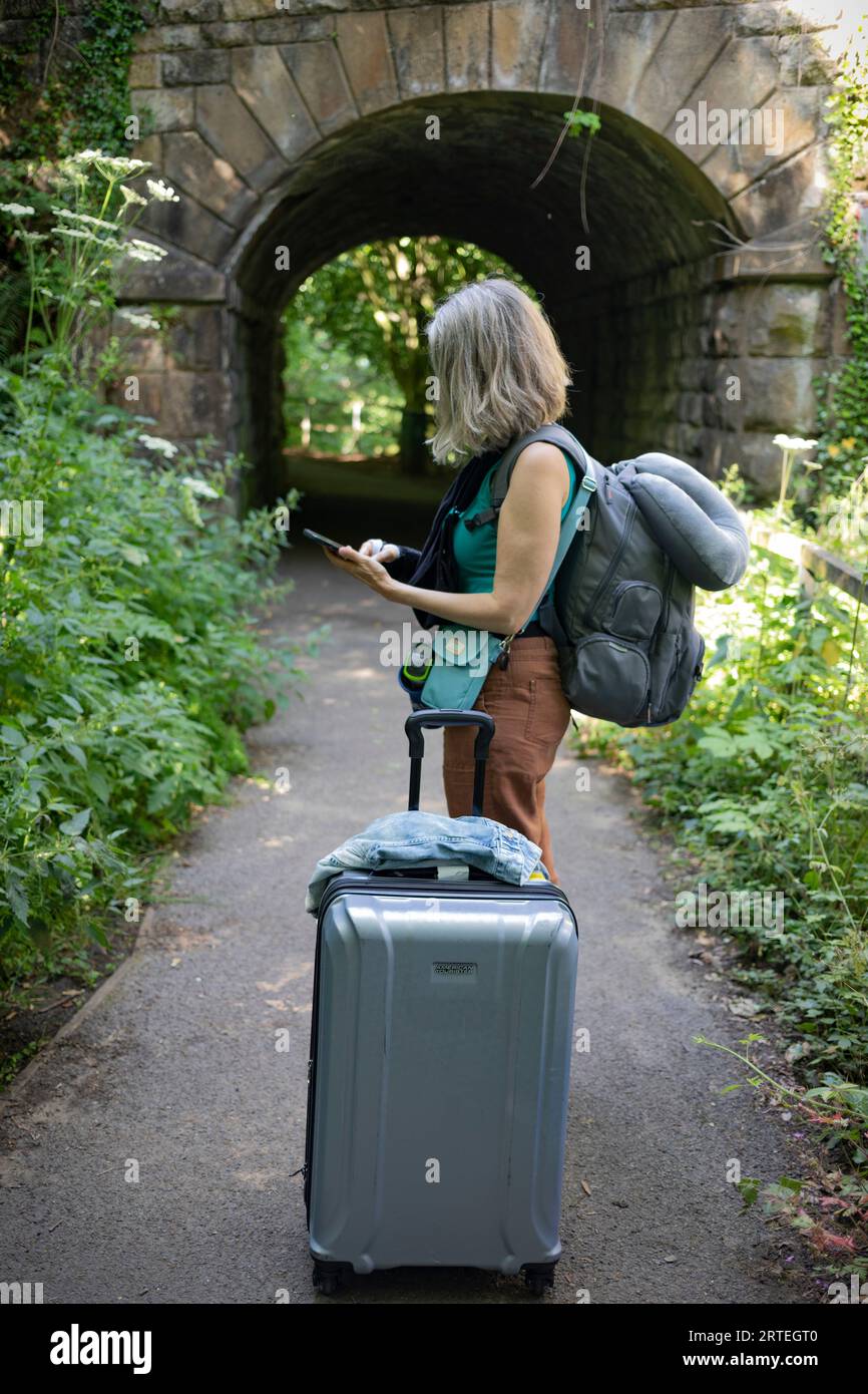 Mature woman uses her smart phone while standing on a path with her luggage; United Kingdom Stock Photo