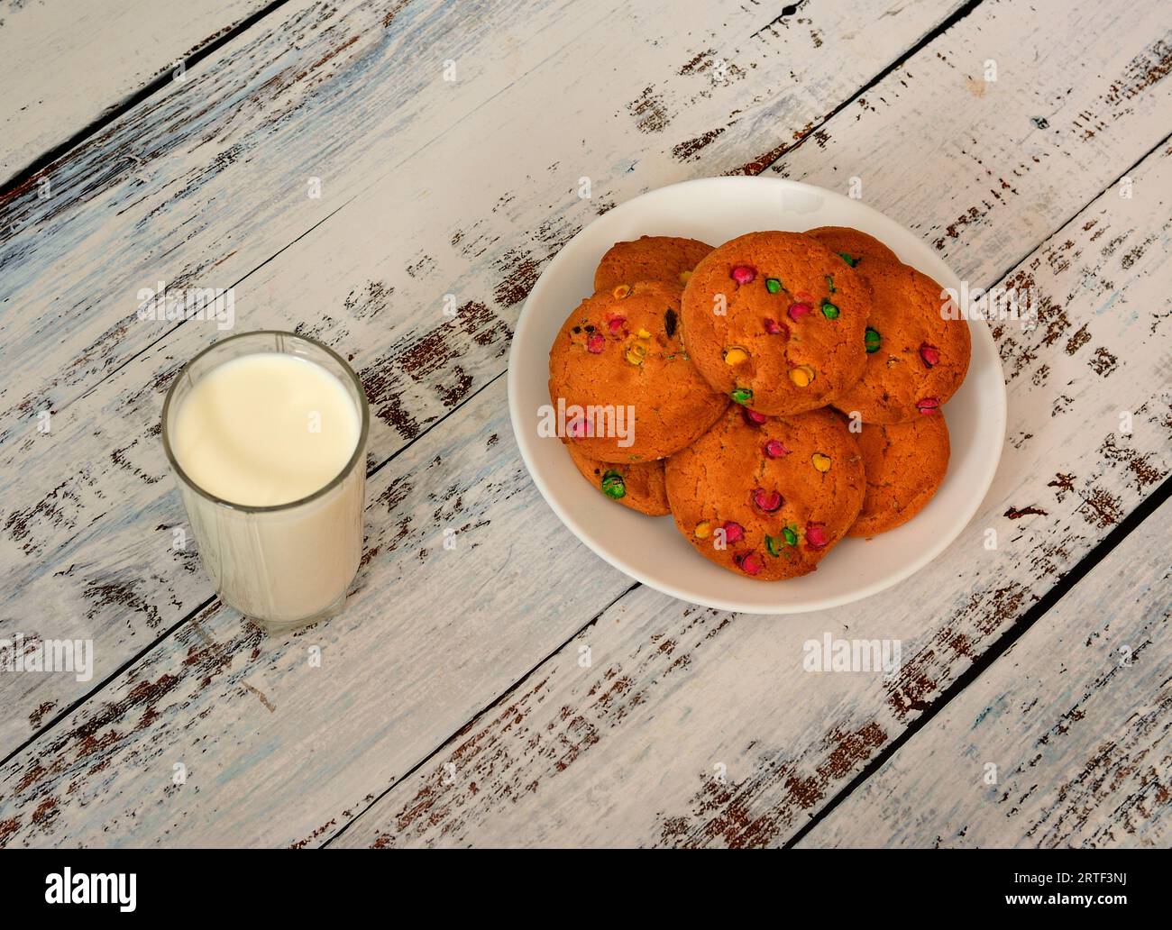 A glass of hot fresh milk and a plate with a bunch of oatmeal cookies with colorful splashes on a light wooden table. Top view, flat lay. Stock Photo
