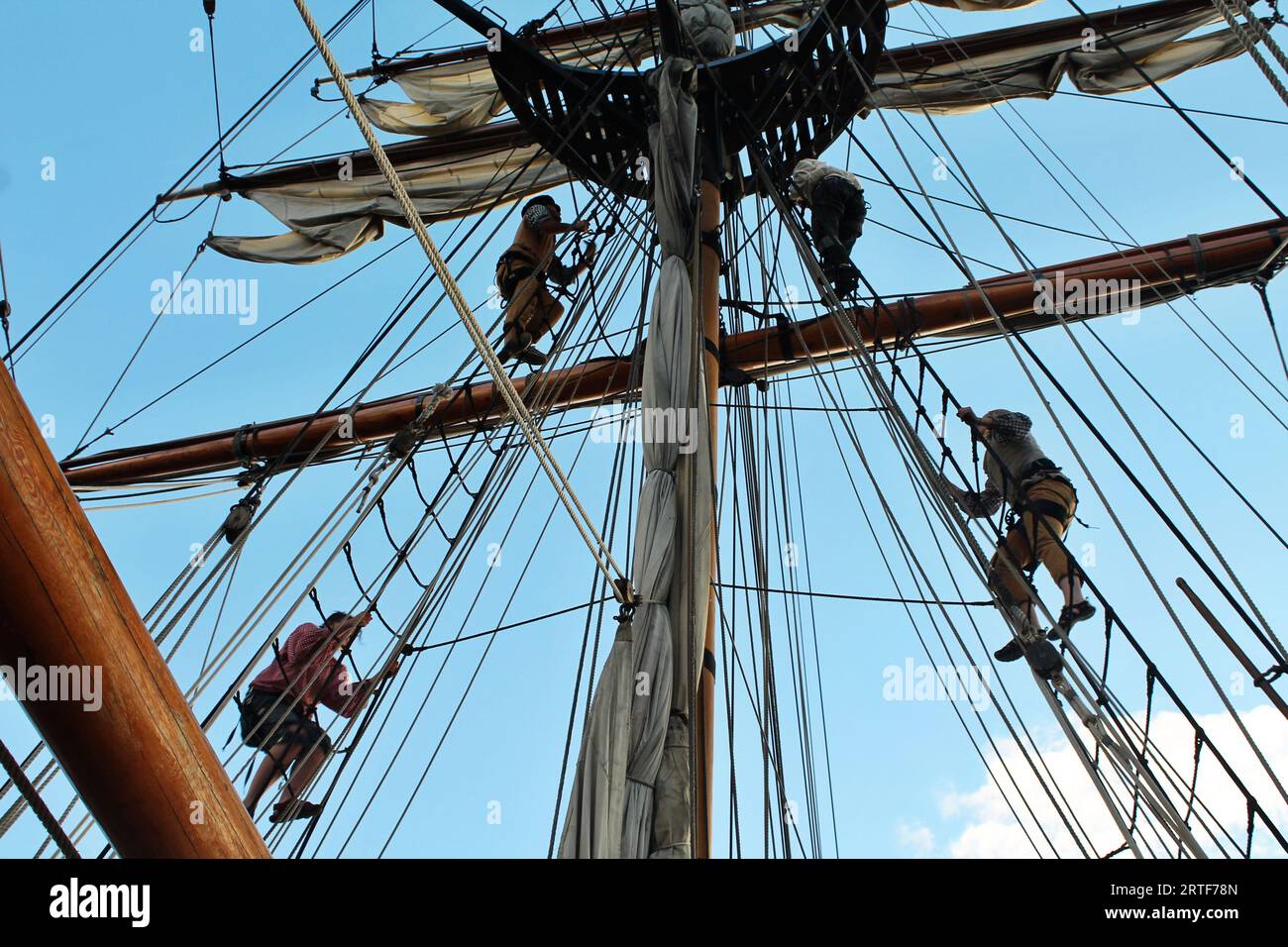 Sailors are seen in silhouette against the blue sky as they climb the rigging of a tall ship to unfurl the sails to catch the rising wind Stock Photo