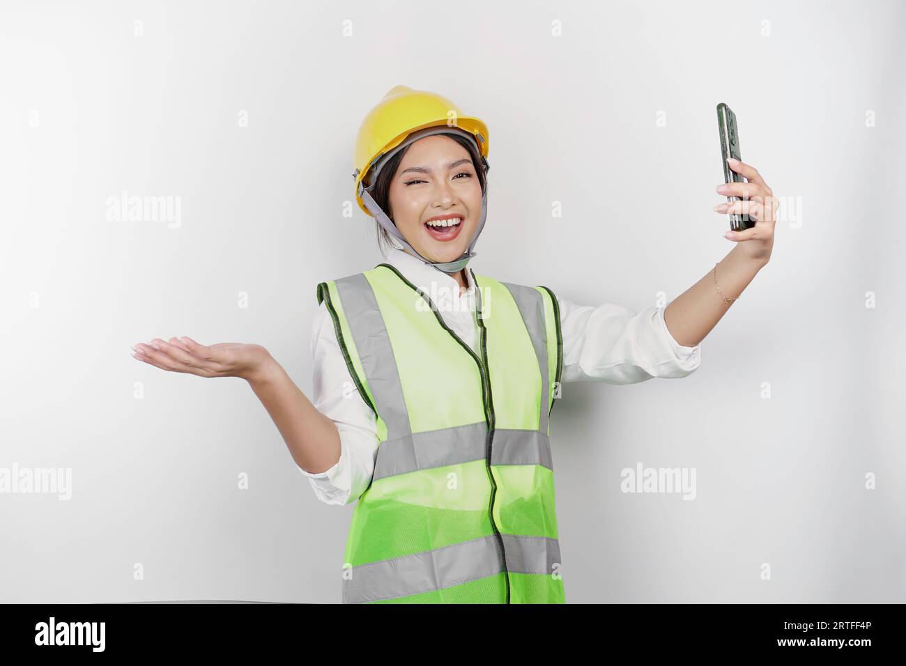 A smiling Asian woman labor wearing safety helmet and vest, holding her phone and pointing to copy space beside her, isolated by white background. Lab Stock Photo