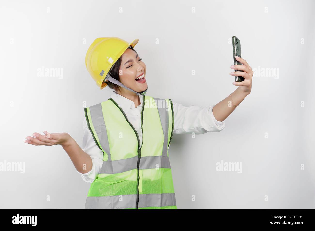 A smiling Asian woman labor wearing safety helmet and vest, holding her phone and pointing to copy space beside her, isolated by white background. Lab Stock Photo