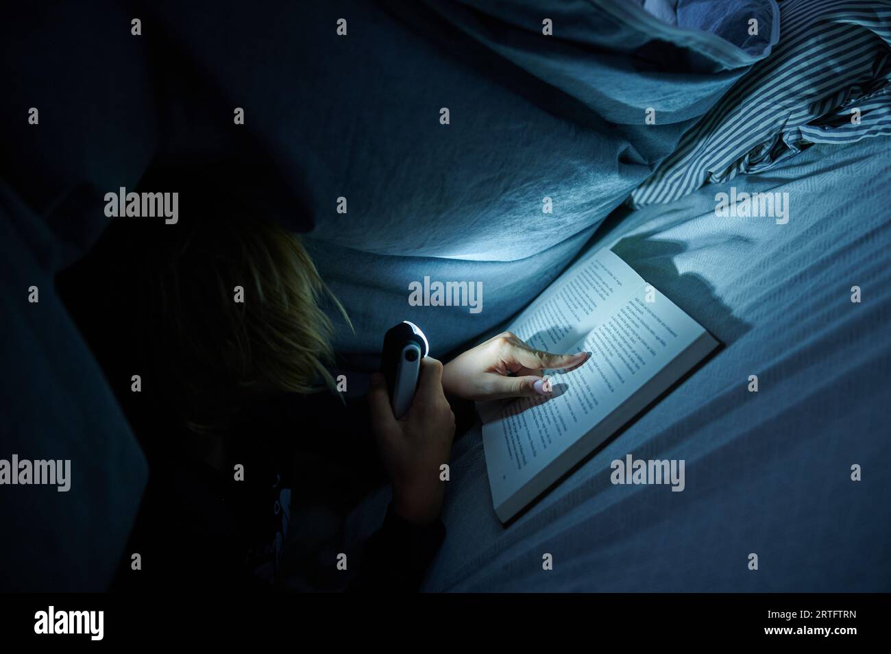 Berlin, Germany. 26th Aug, 2023. A child is lying on a bed in a dark room, reading a book under a blanket with a lamp. (posed scene) Credit: Annette Riedl/dpa/Alamy Live News Stock Photo