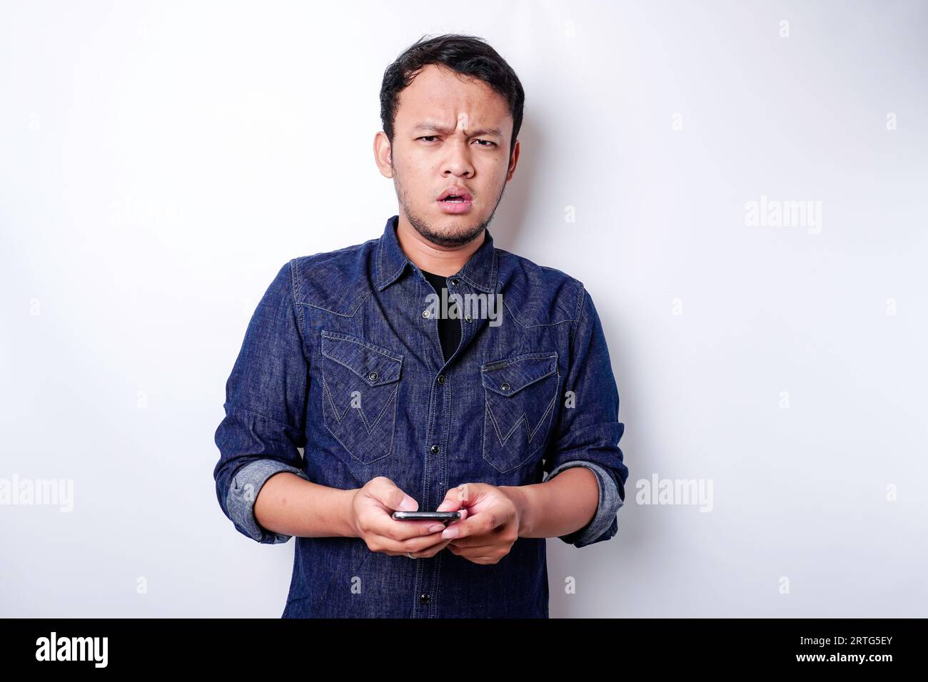 A thoughtful young Asian man is wearing blue shirt holding his phone ...