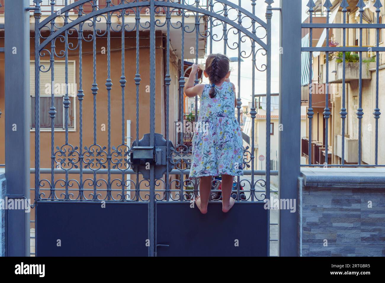 Back view of tanned barefoot little girl wearing blue colorful summer dress climbing wrought iron closed gates, trying to open, waiting, looking. Summ Stock Photo