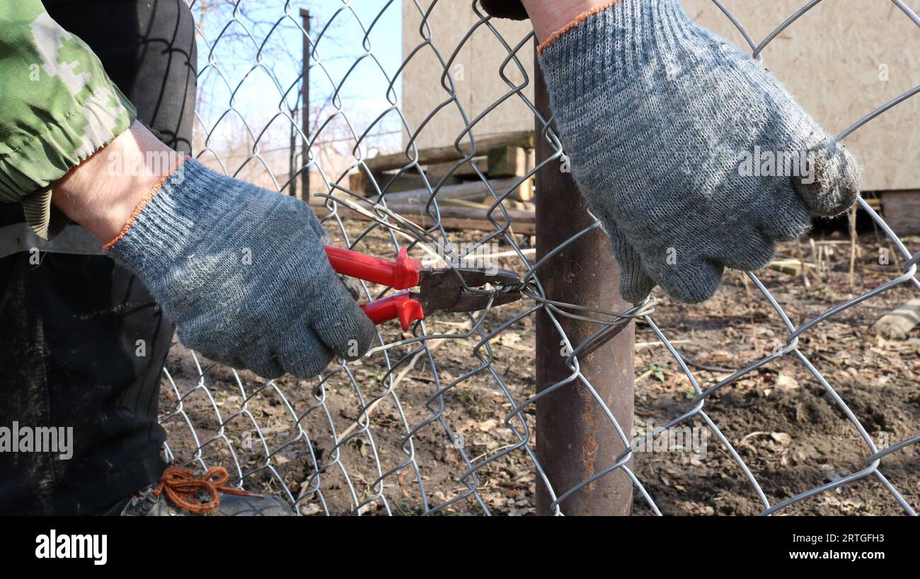 hands in textile gloves with pliers attach galvanized mesh to an iron post using wire in the process of fencing an area on a farm or ranch Stock Photo