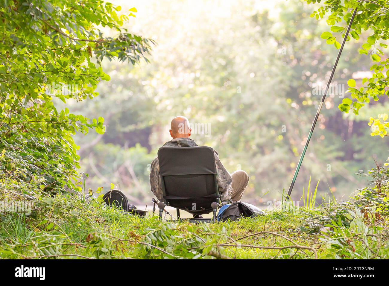 Kidderminster, UK. 13th September, 2023. UK weather: bright sunshine burns through early cloud as summer returns for one happy fisherman today. Rear view of a man fishing at the side of a pool brightly lit by the early morning sunshine. Credit: Lee Hudson/Alamy Live News Stock Photo