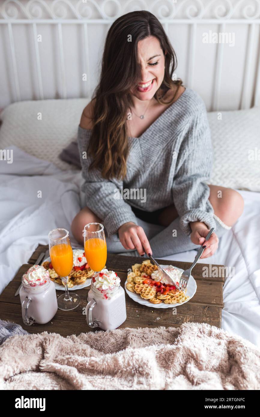 Young woman eating appetizing waffles and tasty drinks on tray on bed Stock Photo