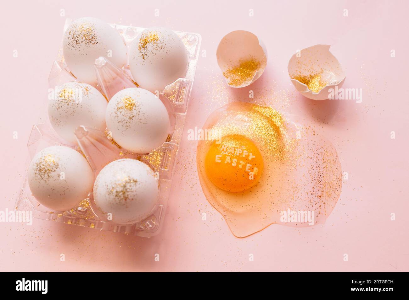 Top view of broken glittered egg with text Happy Easter and raw eggs in plastic box on pink background Stock Photo