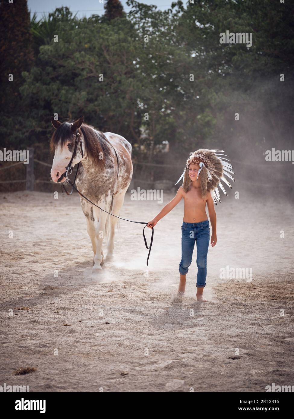 Young slim boy wearing headgear holding reins of horse while strolling on sandy land against green trees and looking away Stock Photo