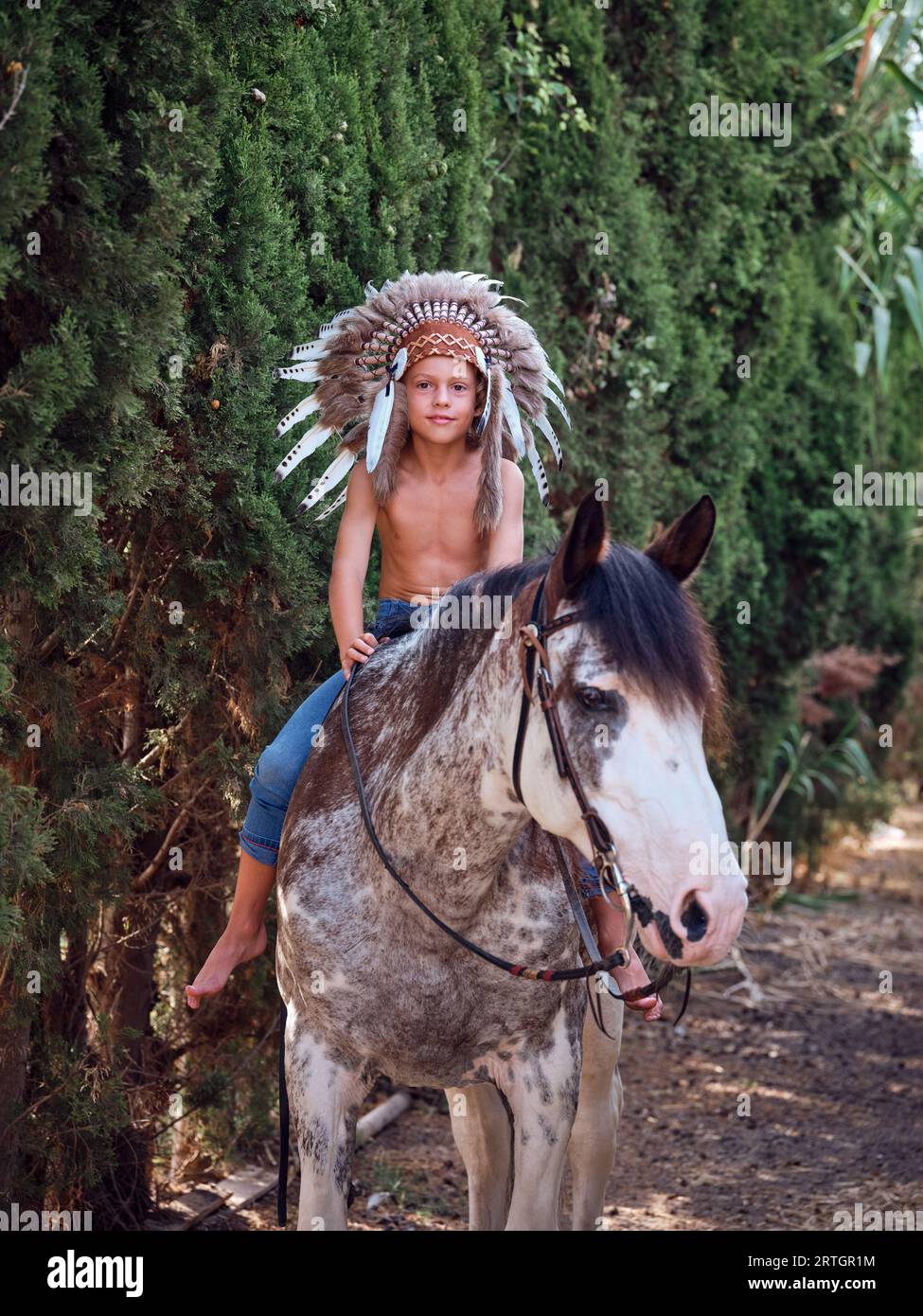 Full body of positive preteen boy in Indian warrior bonnet sitting on horseback and holding reins while riding along path near green trees Stock Photo