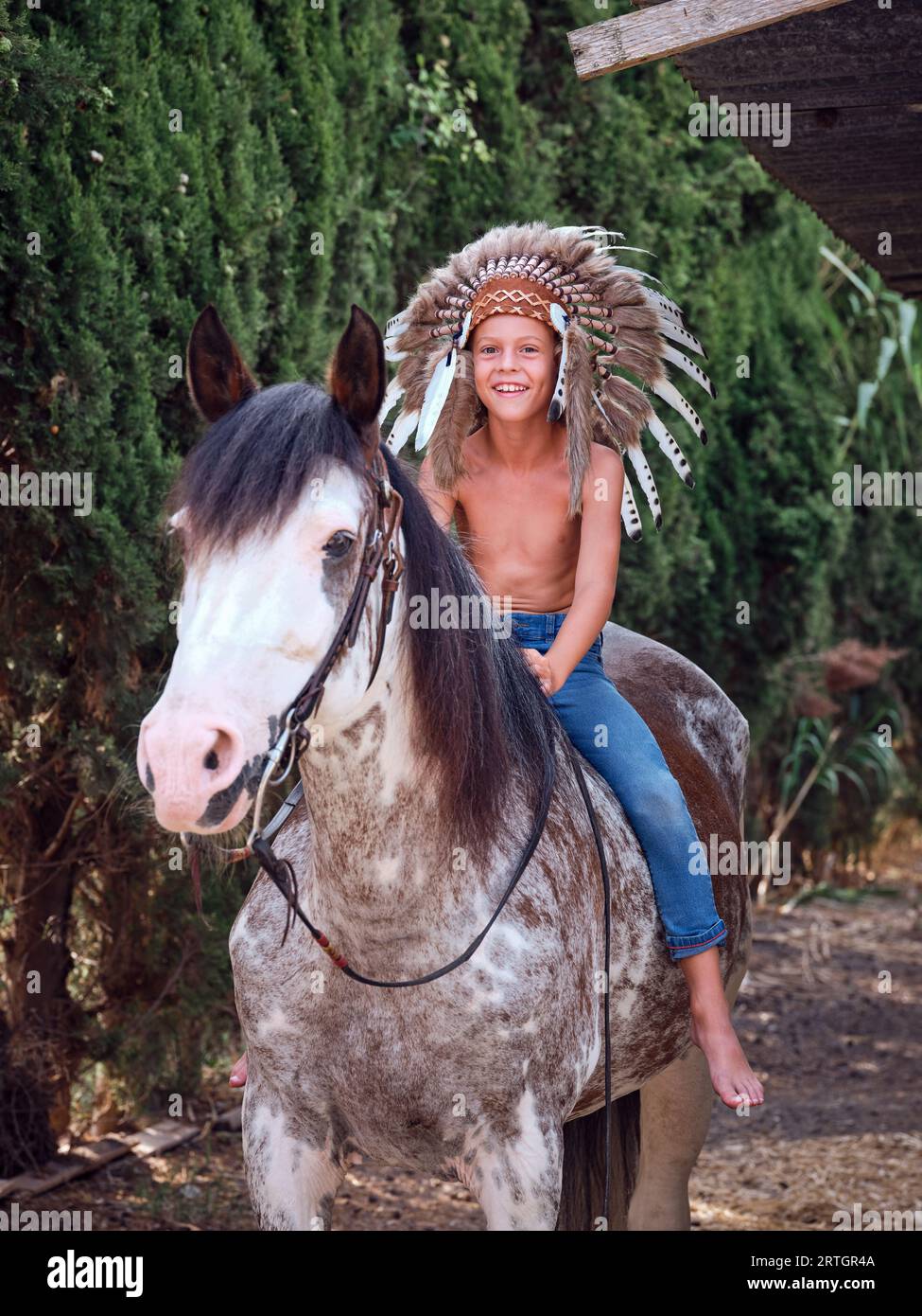 Full body of smiling kid in traditional Indian headwear and jeans sitting on gray horse and holding reins while riding in green park Stock Photo
