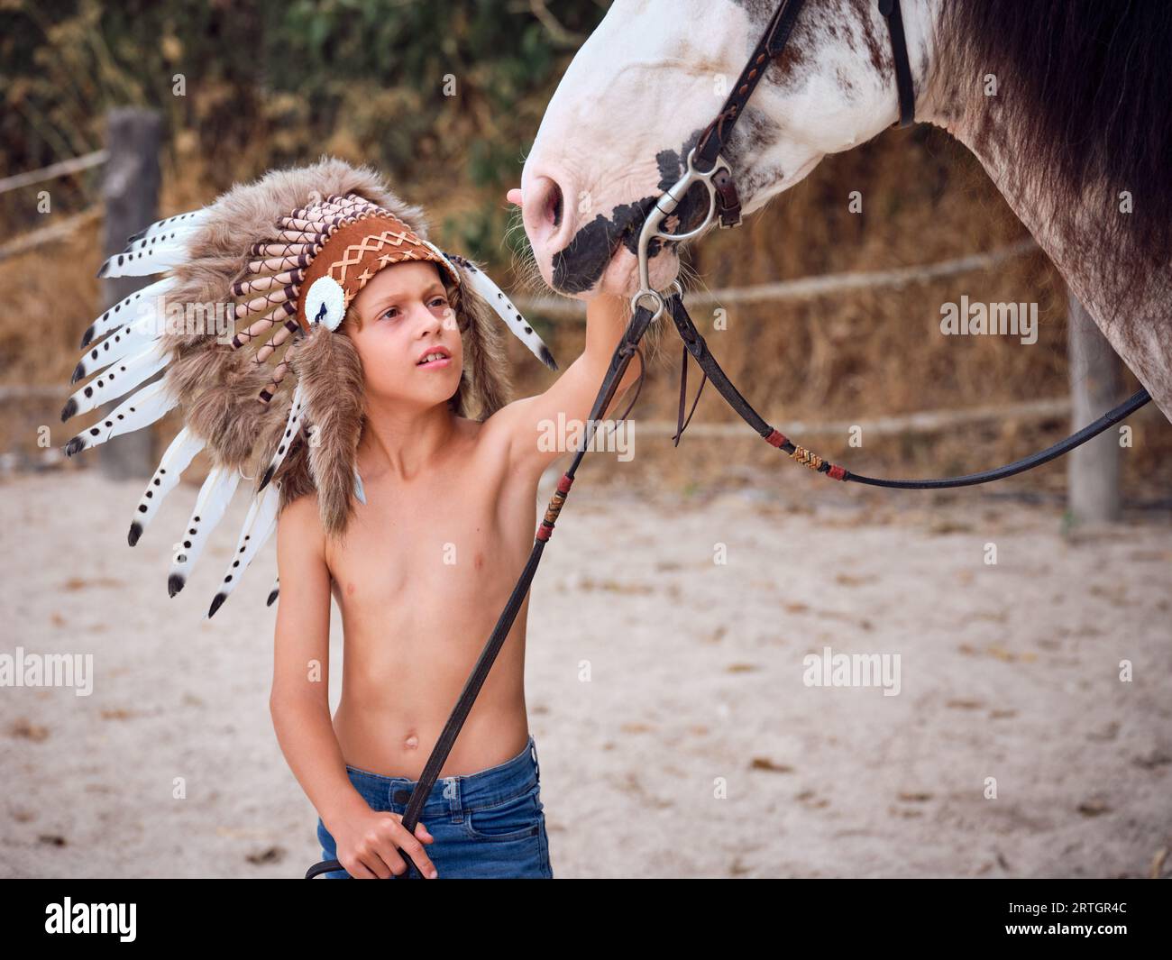 Serious preteen kid in Indian warrior bonnet and jeans standing near horse and holding reins while stroking muzzle in paddock Stock Photo