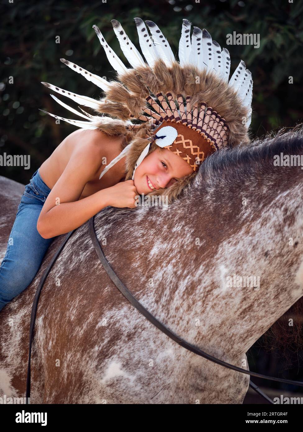 Young boy wearing traditional headwear looking at camera while sitting and hugging horse with reins Stock Photo