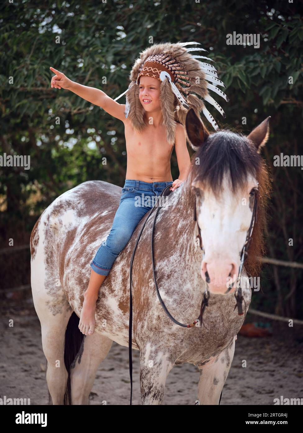 Young boy riding horse in reins while looking away at countryside with green trees and pointing finger to side Stock Photo