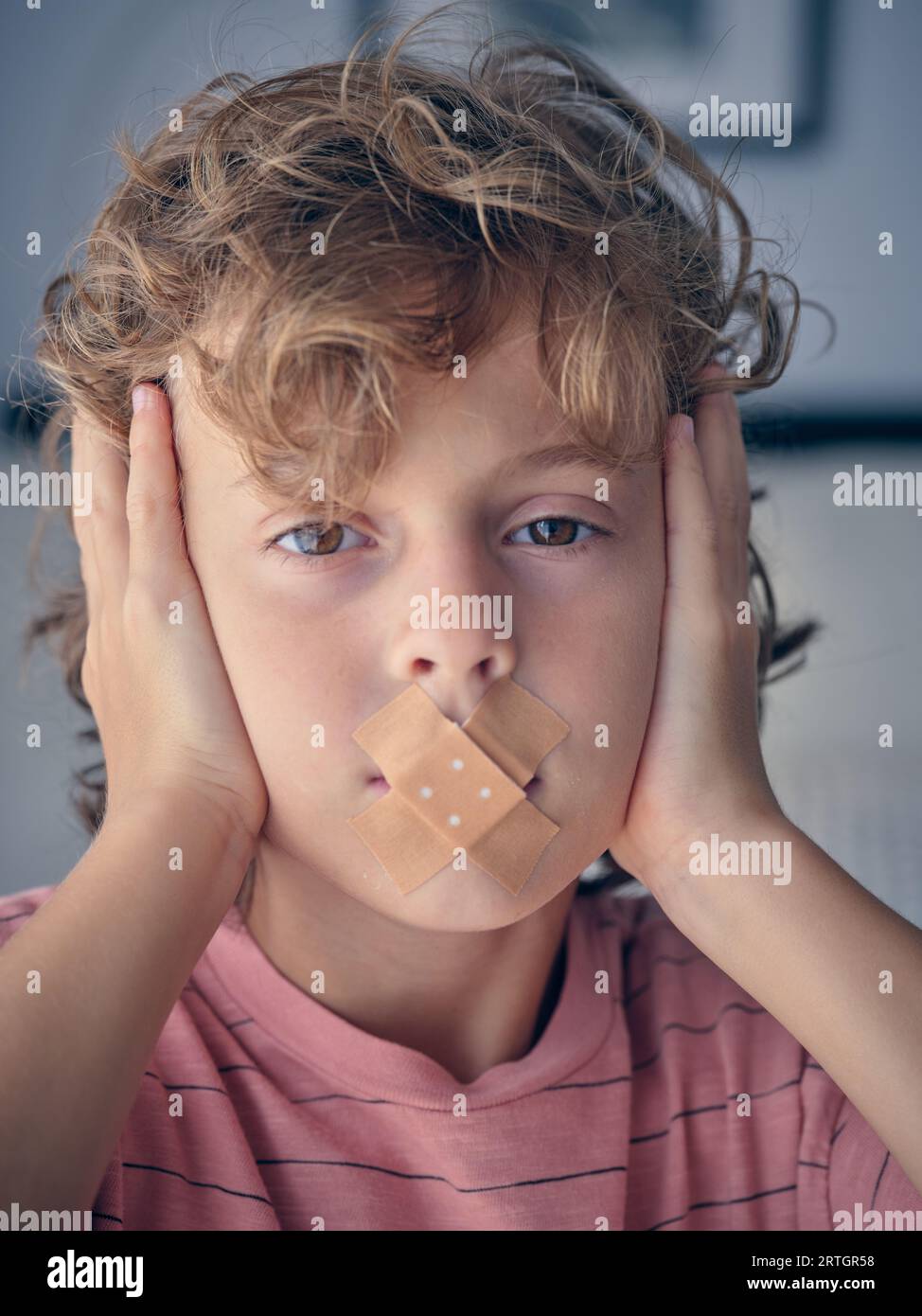 Curly blond haired child with plasters on lips looking at camera while covering ears with hands Stock Photo