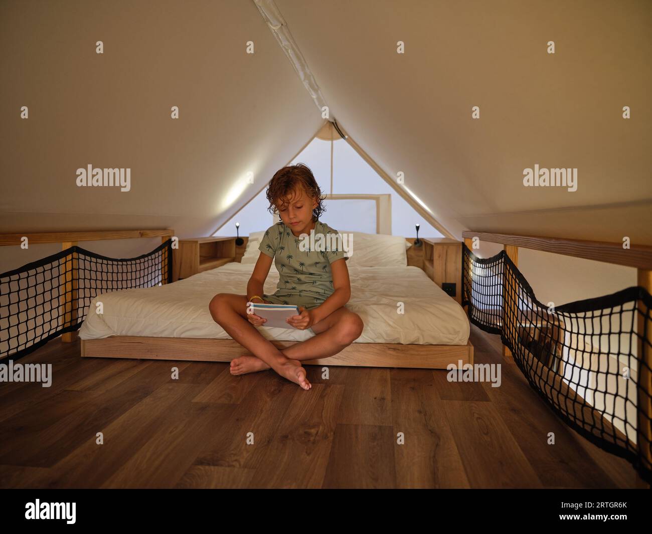 Full body of barefoot child with legs crossed in bedroom holding notebook sitting on comfortable bed under roof of glamping tent Stock Photo