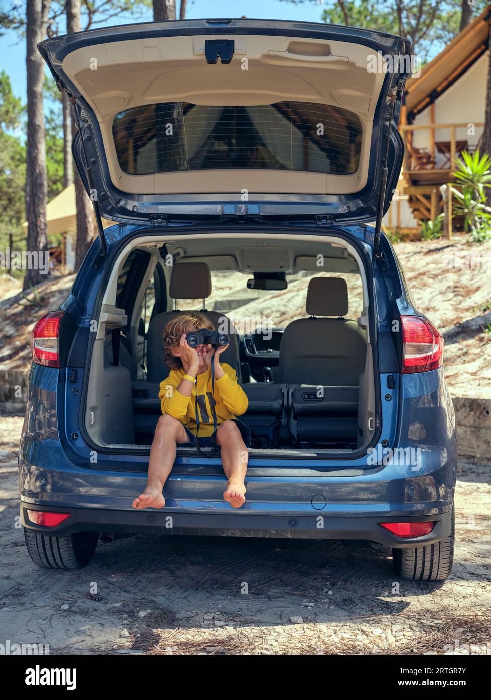 Full body of barefoot child sitting in car trunk near glamping tents in nature and looking away through binoculars while enjoying summer trip Stock Photo