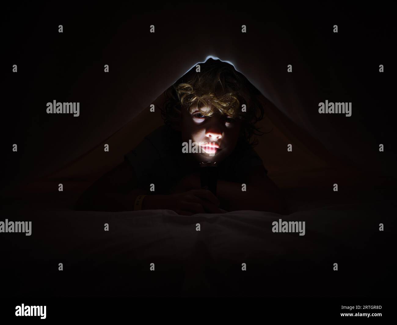 Cute boy illuminating face with bright flashlight while lying on comfortable bed under blanket