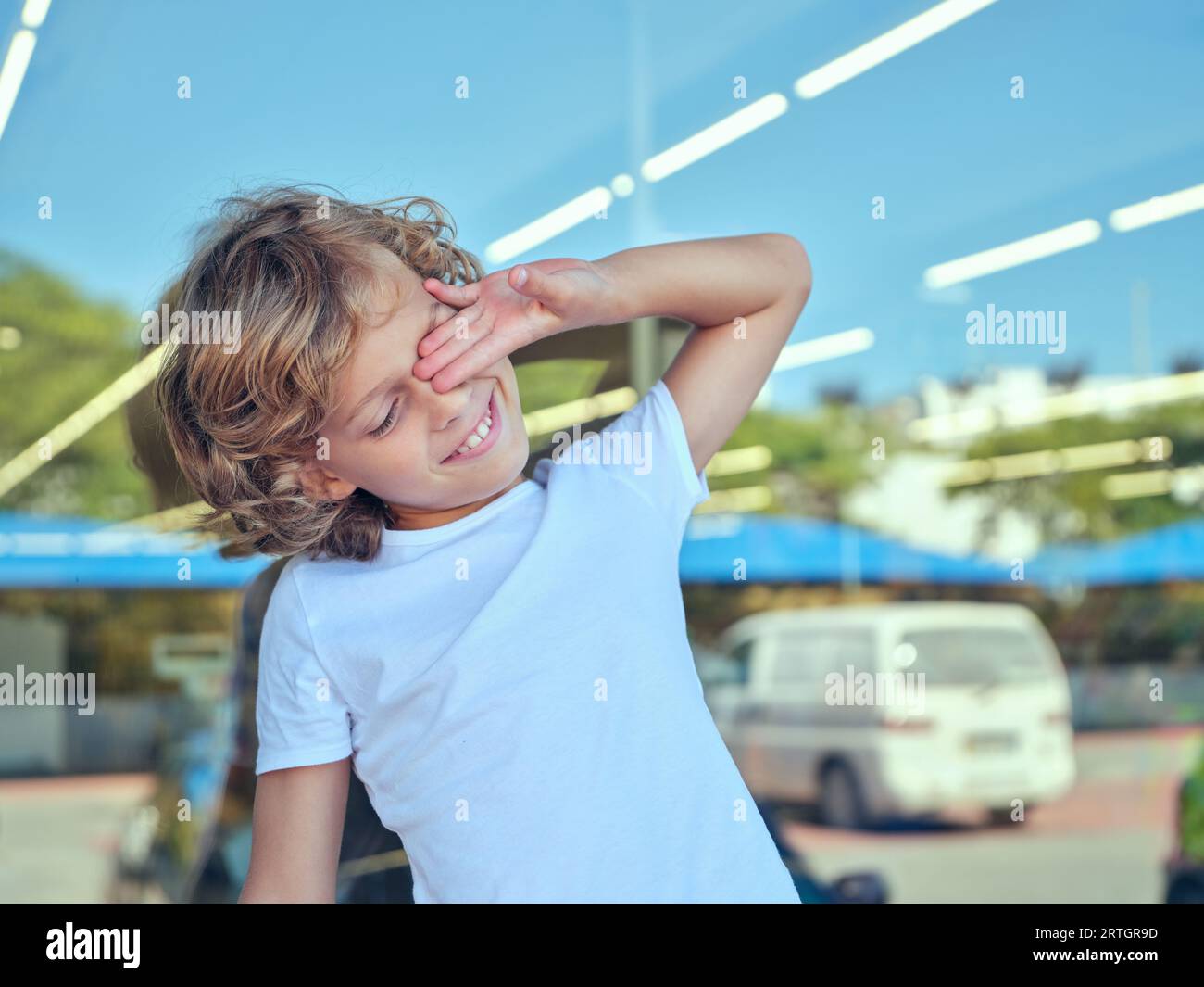Portrait of positive child in casual clothes standing on street against window and rubbing eye while enjoying bright summer day Stock Photo