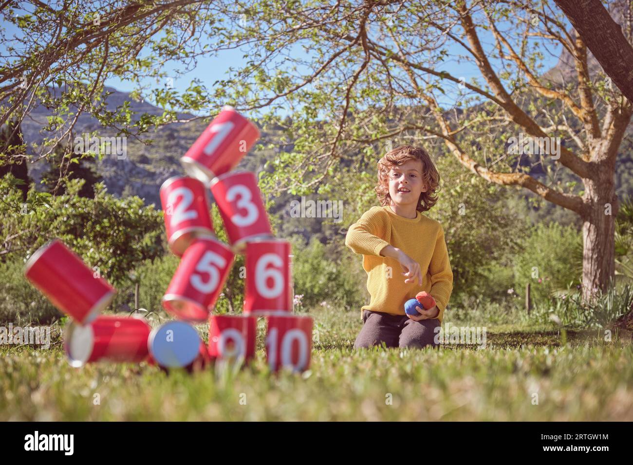 Positive boy kneeling on grassy meadow and throwing bags in pyramid of red tin cans while playing game in nature Stock Photo