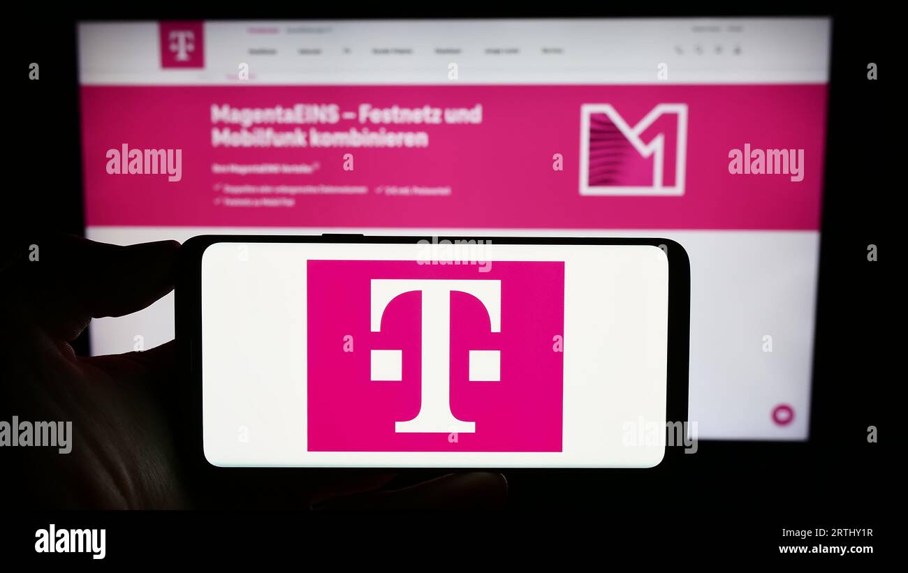 Person holding cellphone with logo of telecommunications company Deutsche Telekom AG on screen in front of webpage. Focus on phone display. Stock Photo