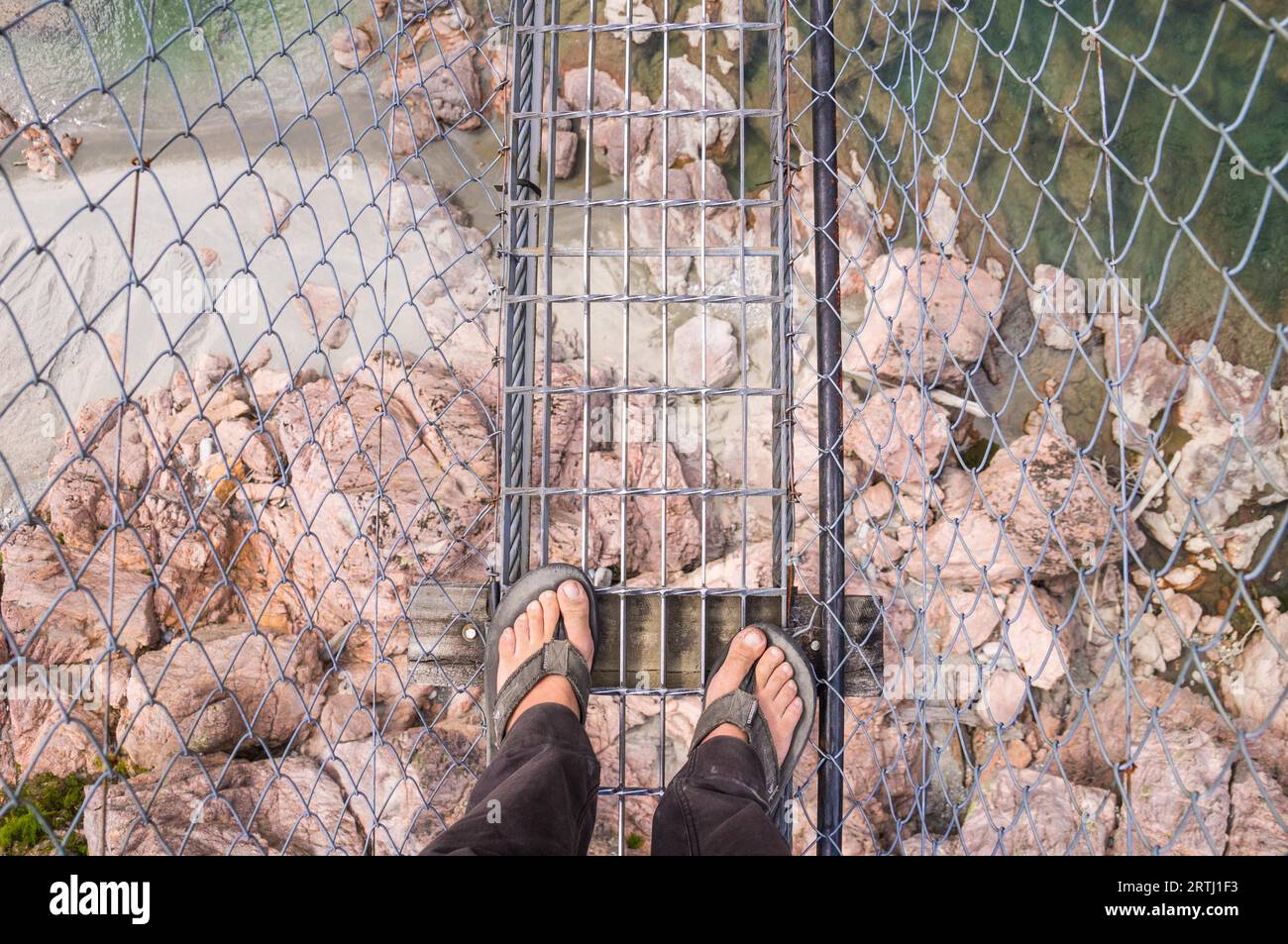 Looking down on feet in flip-flops on a high and dangerous airwalk with a river and rocks below Stock Photo