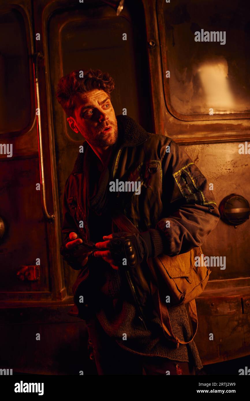 worried man feeling danger and looking away near rusty carriage in post-disaster subway in red light Stock Photo