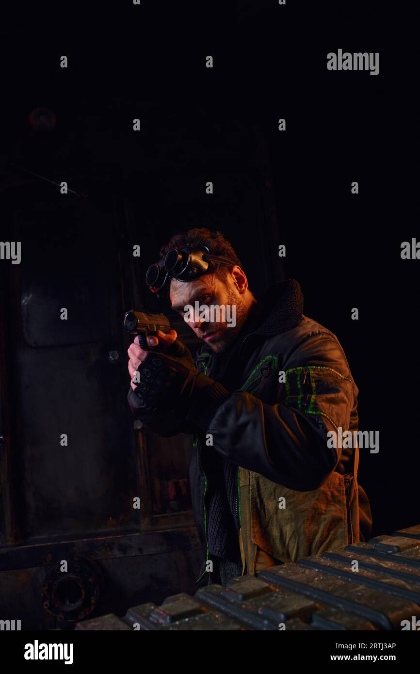 man with military goggles looking away and aiming with gun in red light of post-apocalyptic subway Stock Photo