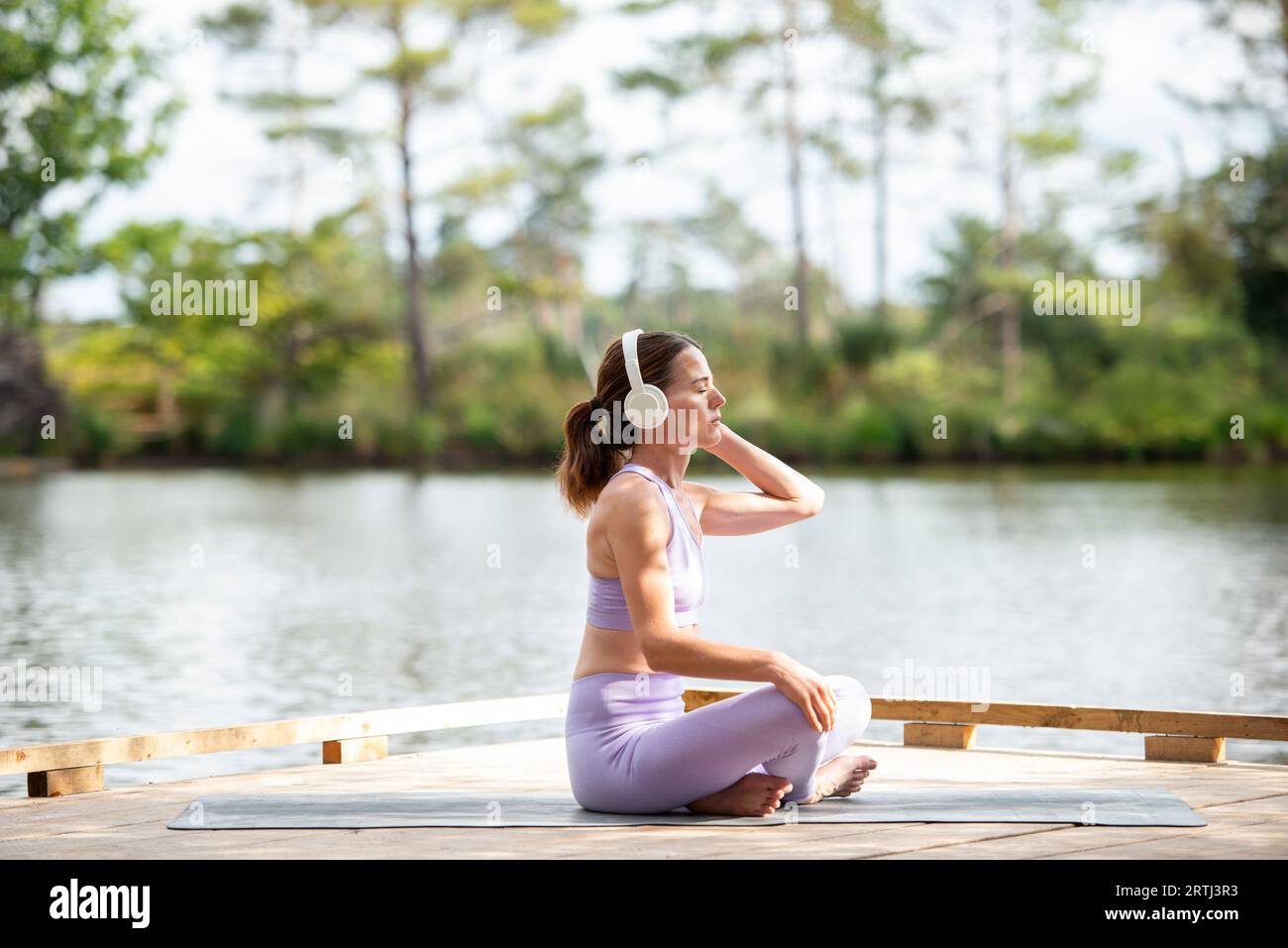 Sporty woman sitting on a jetty by a lake, resting after exercise listening to music on headphones, part of series. Stock Photo