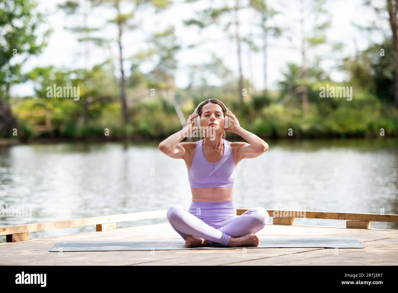 Sporty woman sitting on a jetty by a lake, resting after exercise listening to music on headphones, part of series. Stock Photo