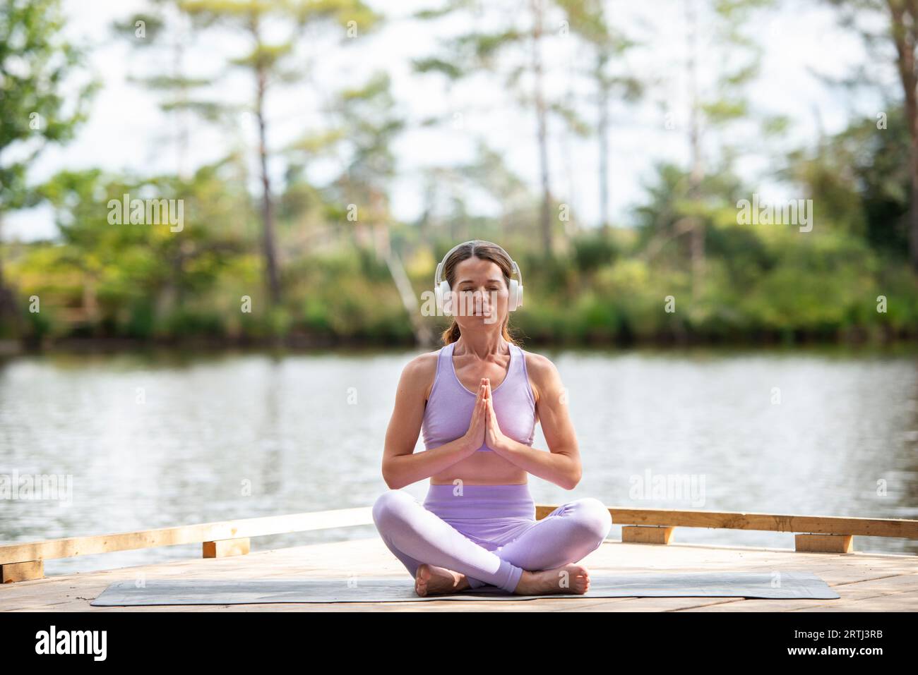 Sporty woman practicing yoga on a jetty by a lake, sitting meditating wearing headphones, part of series. Stock Photo