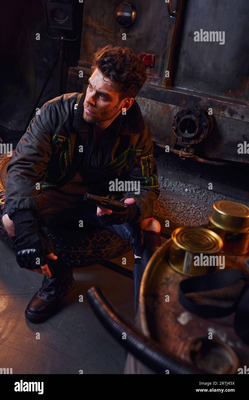 armed man sitting near tins with canned food and looking away in subway, post-apocalyptic survival Stock Photo