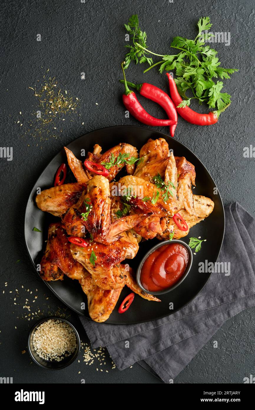 Chicken wings. Grilled or baked chicken wings with sesame seeds and ketchup or spicy tomato sauce on black plate on black or dark slate, stone or conc Stock Photo