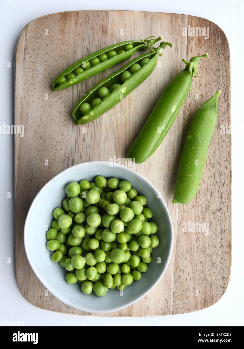 Fresh ingredients on a wooden chopping board with a white background. Garden peas (Pisum sativum) in the pod and in a bowl, taken from above. Stock Photo