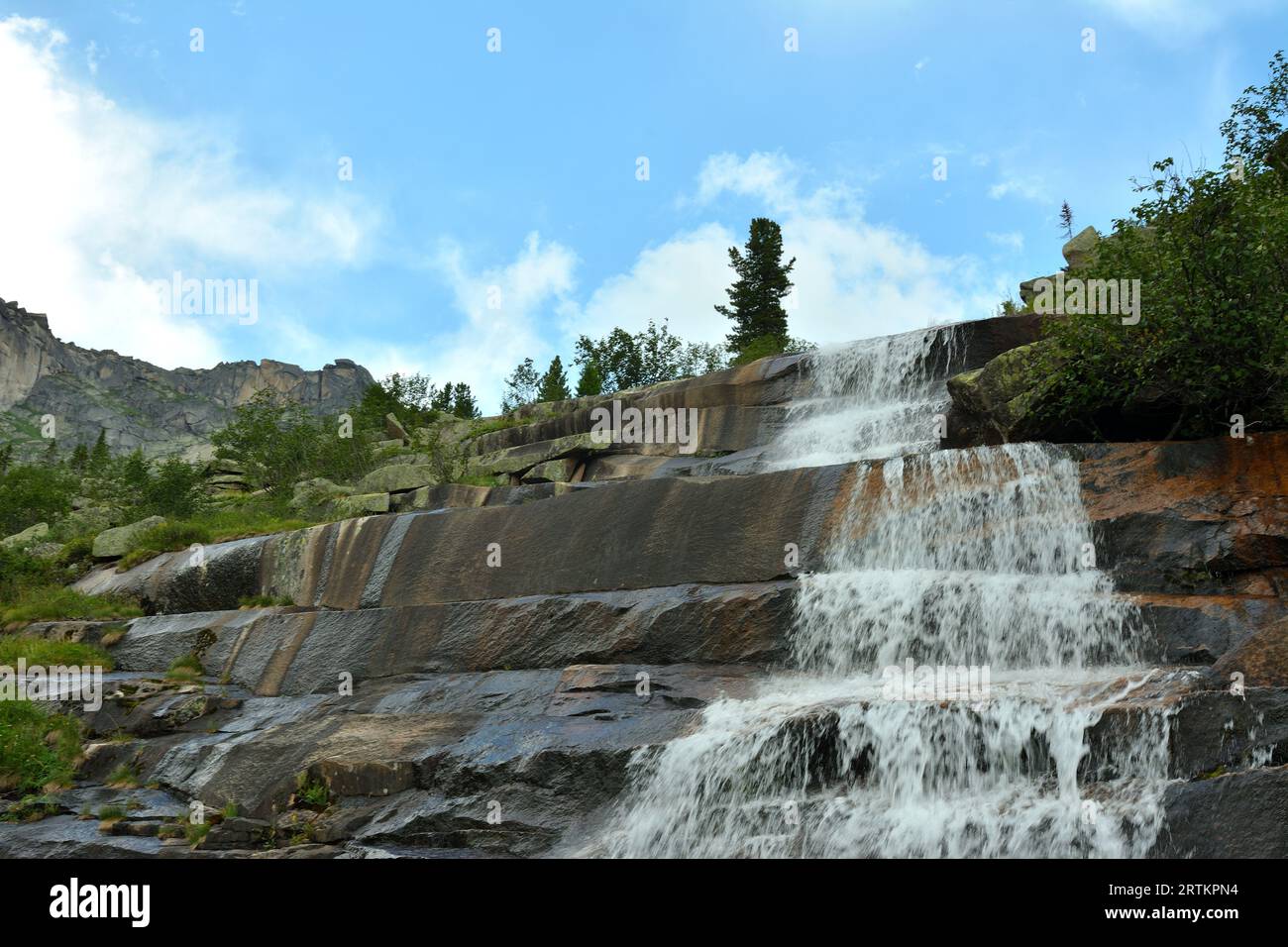 A mountain stream flows in a swift stream from a cascading waterfall in the form of stone steps on a sunny summer day. Jerboa waterfall, Ergaki Nature Stock Photo