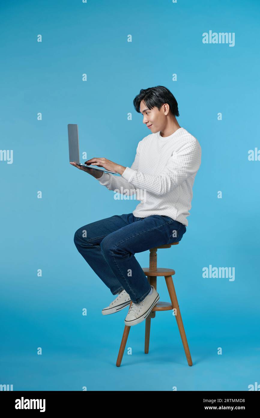 Handsome emotional man sitting on stool and using laptop isolated on blue background Stock Photo