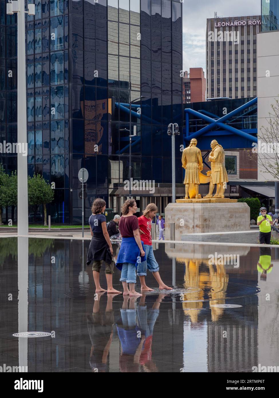 Three girls paddling in the shallow pool Centenary Square, Birmingham, UK; statue of The Golden Boys in the background. Stock Photo