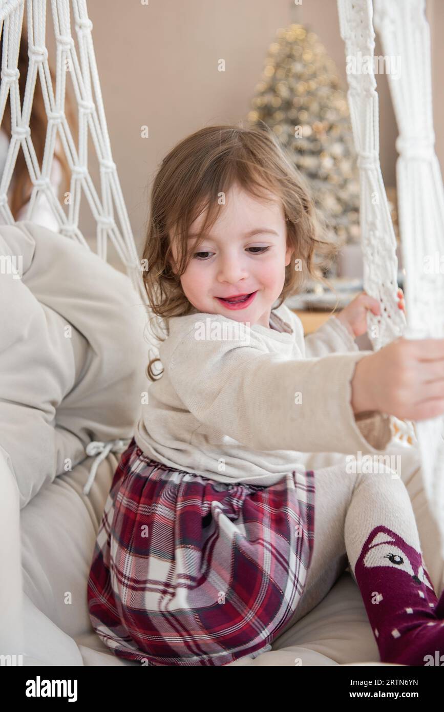 Close-up portrait of funny little girl riding on swing on macrame chair at home. Child swing, playing fooling around. Cozy atmosphere. Rustic style in Stock Photo