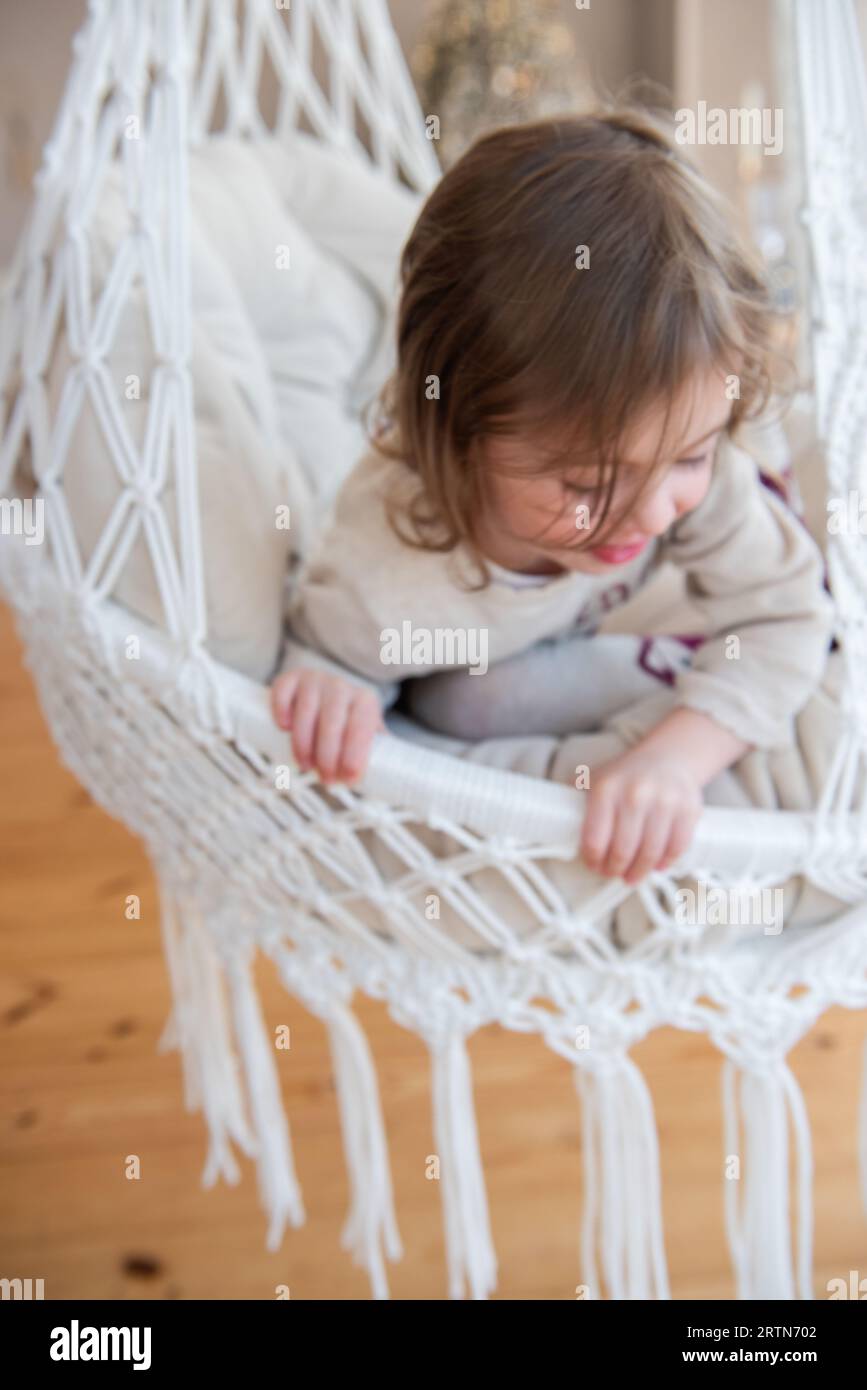 Defocused close-up portrait of funny little girl riding on swing on macrame chair at home. Child swing, playing fooling around. Cozy atmosphere. Rusti Stock Photo