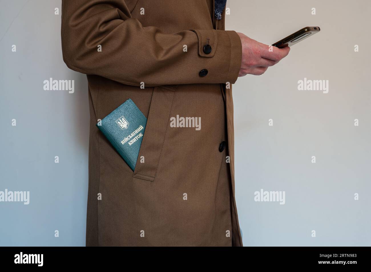 Translation: 'military id'. Civil man holds smartphone in one hand and has ukrainian military id in coat pocket. Mobilization, law, Ukraine Stock Photo